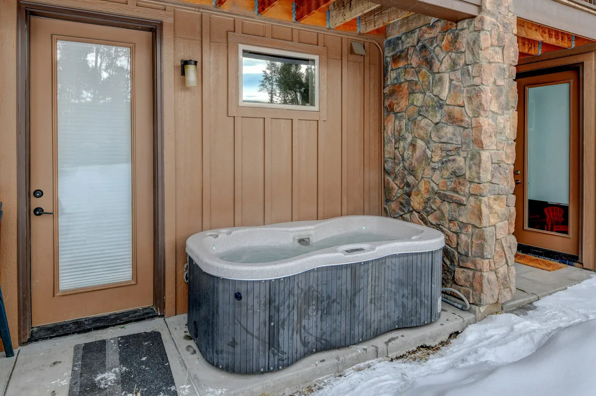 This exterior image showcases a wooden door and stone wall, with a hot tub visible in the foreground surrounded by snow. The scene conveys a welcoming entrance to the villa and emphasizes the natural setting, inviting guests to imagine relaxing outside with their family and pets after a day of mountain exploration.