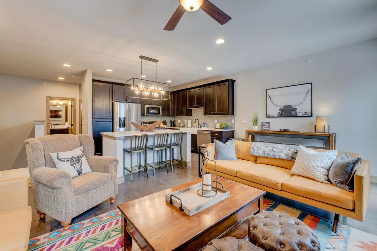 The image showcases the inviting interior of a luxury mountain villa, featuring a stylish open-concept living space with a comfortable seating area, a modern kitchen island, a vibrant area rug, and tasteful wall art. The scene exudes warmth and comfort, ideal for socializing, cooking, relaxing, and entertaining guests.