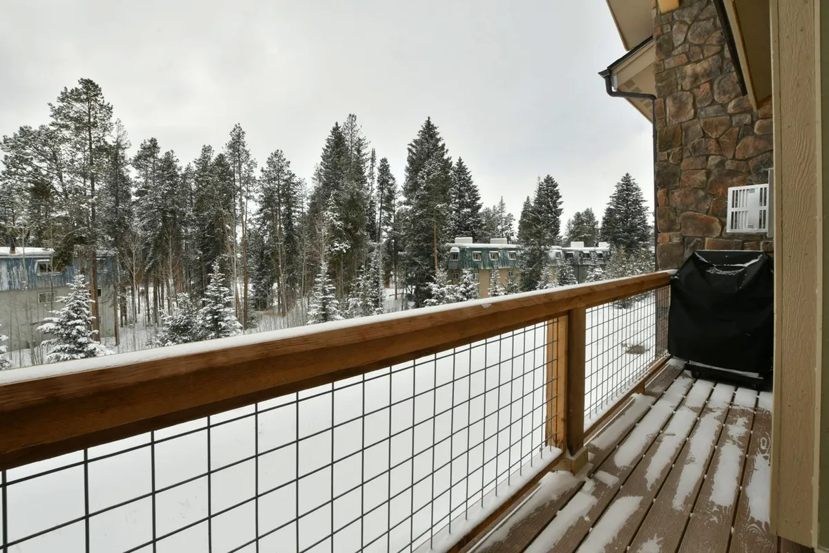 A wooden porch railing overlooks a tranquil, snow-covered landscape dotted with tall evergreen trees. Guests can imagine relaxing outside, taking in views of the winter wonderland, or even enjoying evenings around a fire pit. The serene setting captures the essence of mountain living and the inviting outdoor spaces that complement the villa experience.