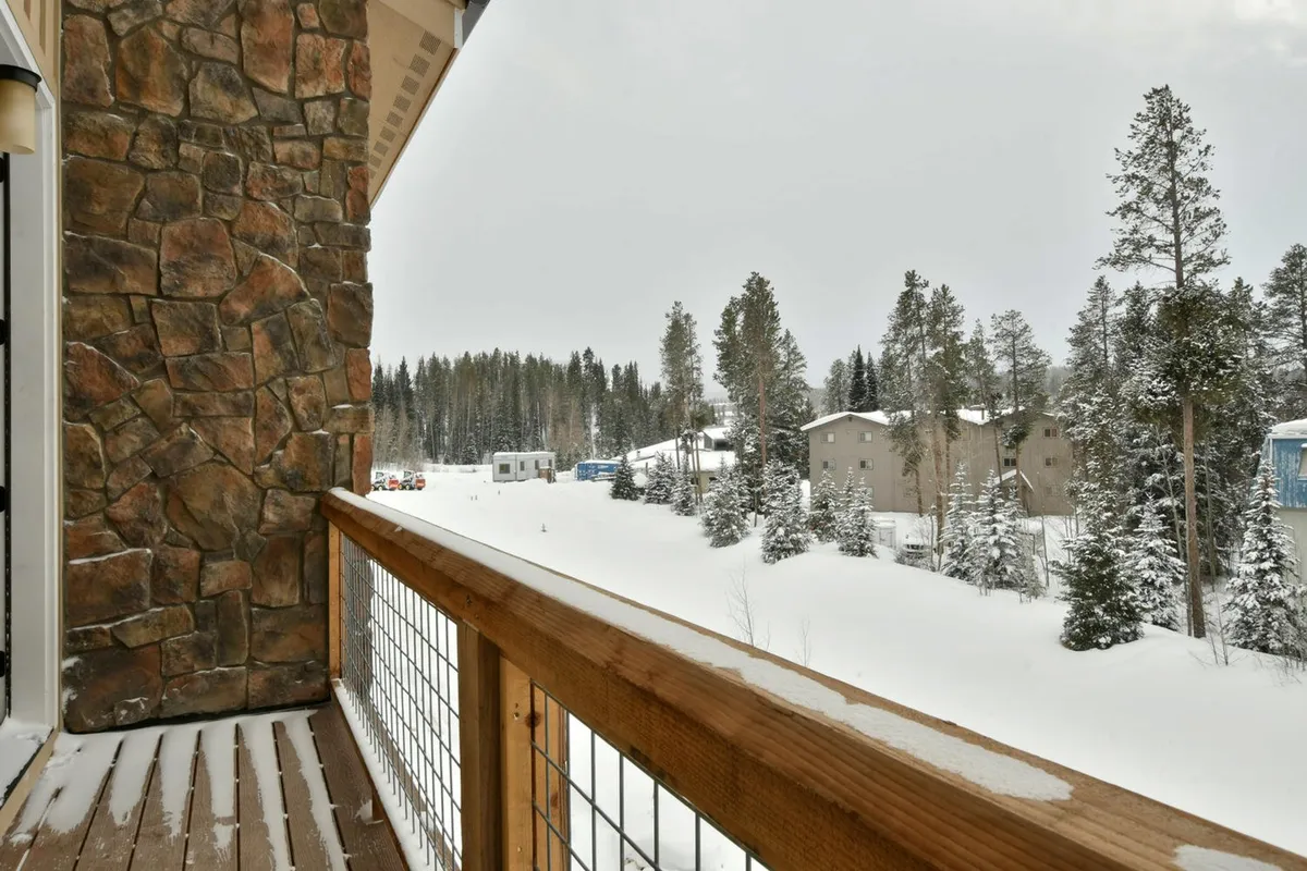 A snowy, tranquil landscape with a stone wall in the foreground, pine trees dusted with fresh snow, and distant houses nestled in the background. The scene invites thoughts of winter adventures, with fresh powder perfect for skiing, snowboarding, or simply exploring nature trails right outside.