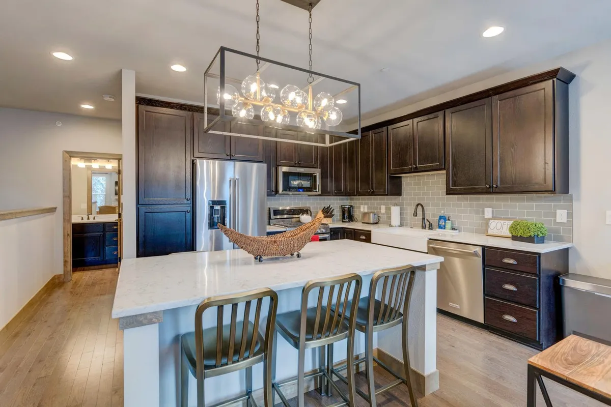 A close perspective of the kitchen island, highlighting elegant pendant lighting, dark cabinetry, and stainless steel appliances. The setup suggests a space designed for both casual brunches and more formal dinner parties, with ample prep and serving space.