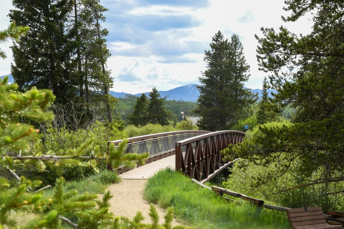 A scenic wooden bridge nestled among trees with a stunning mountain backdrop, inviting exploration. The natural setting suggests access to nearby walking trails and opportunities for nature observation and peaceful strolls.