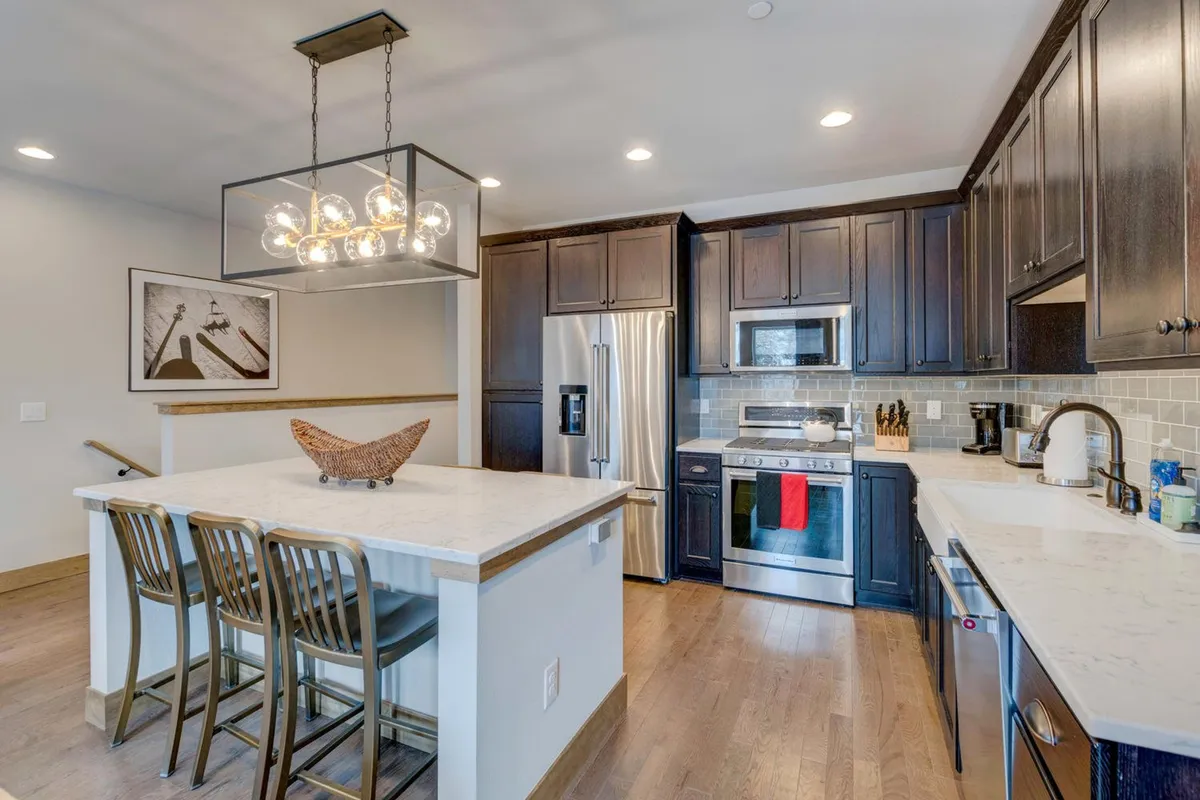 An inviting view of the kitchen featuring a prominent kitchen island beneath a striking chandelier, surrounded by dark cabinetry and gleaming stainless steel appliances. The space is bustling with the energy of cooking and entertaining, and there's ample room for multiple people to participate in meal preparation or conversation.