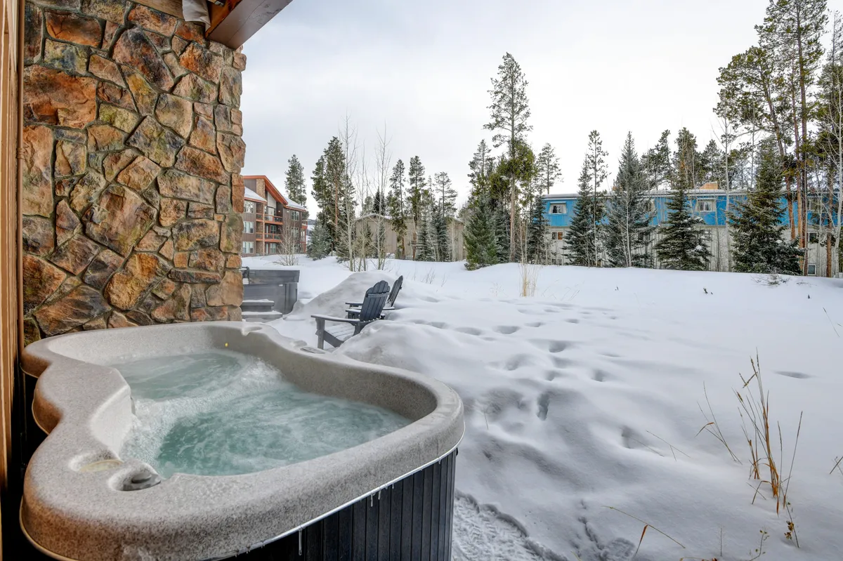 A pristine outdoor hot tub is set against a snowy, pine-lined landscape. The inviting warm waters of the tub steam gently in the crisp mountain air, while a backdrop of snow-laden trees and an expansive sky suggest seclusion and relaxation. The space feels private and luxurious, ideal for unwinding after a day of mountain adventures or enjoying stargazing at night.