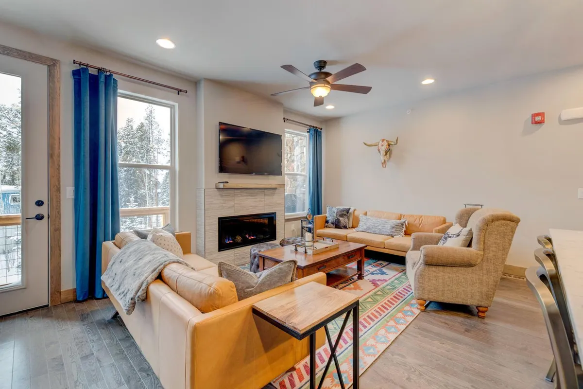 A spacious great room featuring a contemporary gas fireplace as its centerpiece, surrounded by plush sofas and a cozy rug. A large coffee table sits in the center, inviting gatherings for games or conversation. The wide windows flood the room with natural light and offer glimpses of the stunning mountain landscape. A smart TV is mounted near the fireplace, blending entertainment with comfort.