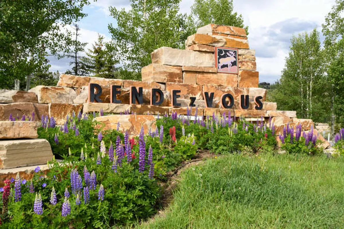 A welcoming exterior shot featuring the 'RENDEZVOUS' signage set amidst lush flower beds and a natural stone structure. The garden and inviting entrance suggest a vibrant, community-oriented atmosphere and connect visitors to nearby trails and local activities.
